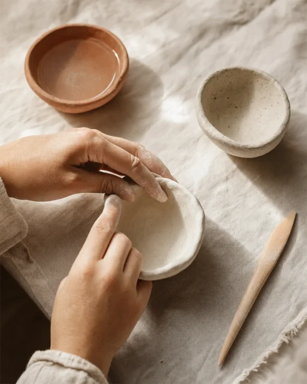 Hands pinching the wall of a small white air dry clay bowl on pale linen